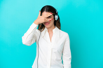 Telemarketer Italian woman working with a headset isolated on blue background covering eyes by hands and smiling