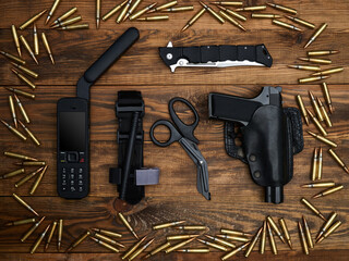 Close-up on a wooden background. Satellite phone with a large antenna, tactical knife, handgun, atraumatic scissors, and hemostatic tourniquet surrounded by a frame of cartridges - obrazy, fototapety, plakaty