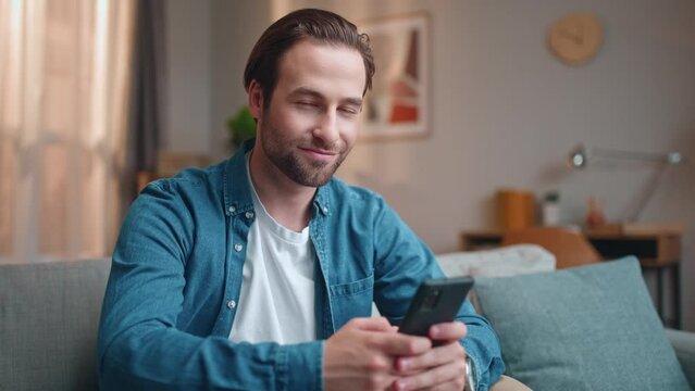 Cheerful man sitting on sofa and using smartphone. Satisfied male sitting on couch. Happy man rising head and smiling at camera. Male holding gadget and scrolling social media.
