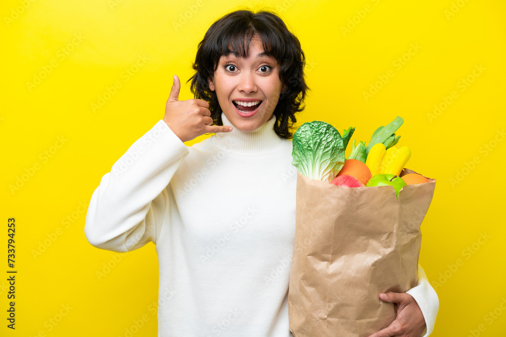 Wall mural young argentinian woman holding a grocery shopping bag isolated on yellow background making phone ge