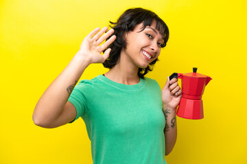 Young Argentinian woman holding coffee pot isolated on yellow background saluting with hand with...