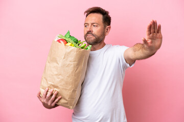 Middle age man holding a grocery shopping bag isolated on pink background making stop gesture and...
