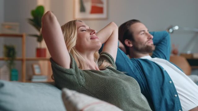 Spouses sitting on couch. Man and woman putting hands above head. Smiling male and female relaxing at apartment. Man and woman sitting on sofa. Cute couple spending weekend together.