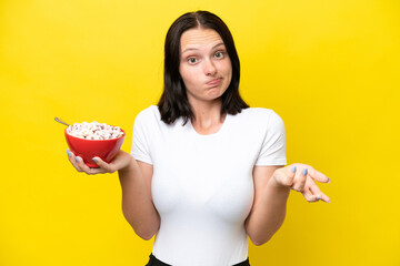 Young caucasian woman holding a bowl of cereals isolated on yellow background making doubts gesture while lifting the shoulders