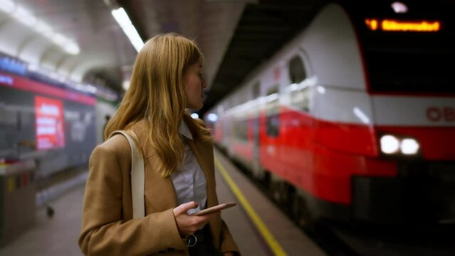 Female standing at subway platform with mobile phone, using social media and waiting train