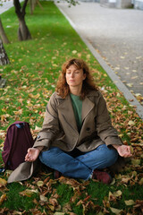 Caucasian middle aged woman meditating in lotus pose at autumn city street with leaves. Woman relaxing on yellow leaves. Meditation, Mental health, self care, mindfulness. High quality photo