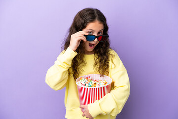Little caucasian girl isolated on purple background surprised with 3d glasses and holding a big bucket of popcorns