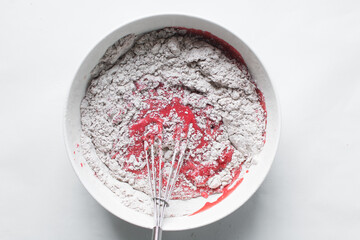 Top view of flour being folded into Red velvet cake batter in white ceramic mixing bowl, process of...