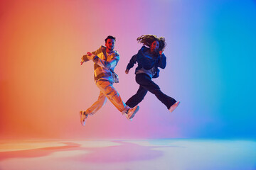 Two dancers in mid-air jump while dancing in motion against gradient blue-orange background in neon...