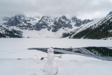 Cute snowman against a frozen lake Morske Oko and rocky mountains background, Tatra national park, Poland. Scenic winter or early spring landscape, travel and hiking adventure concept