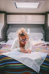 One woman planning next travel trip destination laying in bed inside a mpodern camper van motorhome. People and vanlife lifestyle traveler. Female looking paper map guide for road map indoor leisure