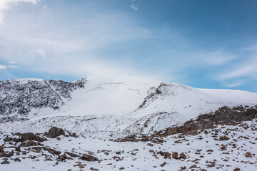 Cold snow-white landscape with snow-capped peak of dome shape under clouds in blue sky. Snow-covered rocky mountain range with snowy dome shaped mountain top. Pure white snow and stones in mountains.