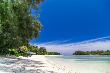 Tropical bay, paradise destination on the Cook Islands. Rarotonga tropical beach with umbrellas and sunbeds. Coast with palm trees during a sunny day. Blue sky with clouds and turquoise water.
