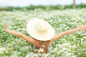 Back view of a little smiling girl in a blue dress and a straw hat on a chamomile field on a sunny summer day