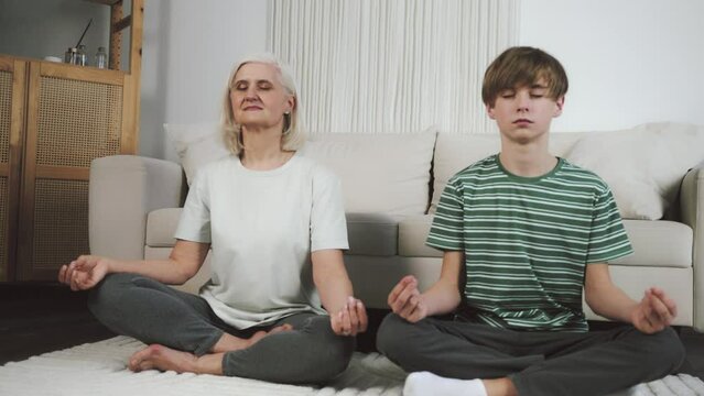 White-haired senior woman and teenaged boy meditating, doing yoga, breathing deeply, practicing mindfullness. Active family staying at home in a big living room, listening to silence, crearing minds
