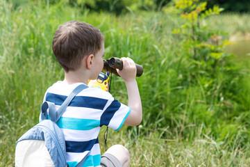 Little boy with binocular and backpack looking for the right path in summer park outdoor. Child exploring forest nature and environment on sunny day during outdoor ecology