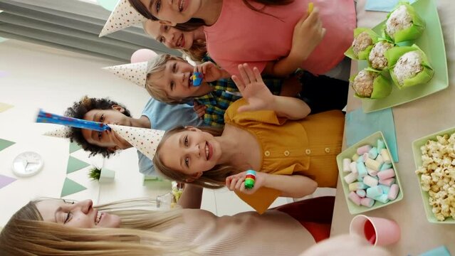 Vertical POV of Caucasian woman filming selfie video with diverse school children in party hands standing by table with sweets on, using blow ticklers, waving, smiling, looking on camera Vertical POV 