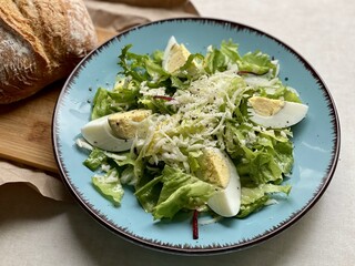 Green lettuce leaves on a plate, garnished with slices of egg and cheese with a bun on a cutting board next to it.
