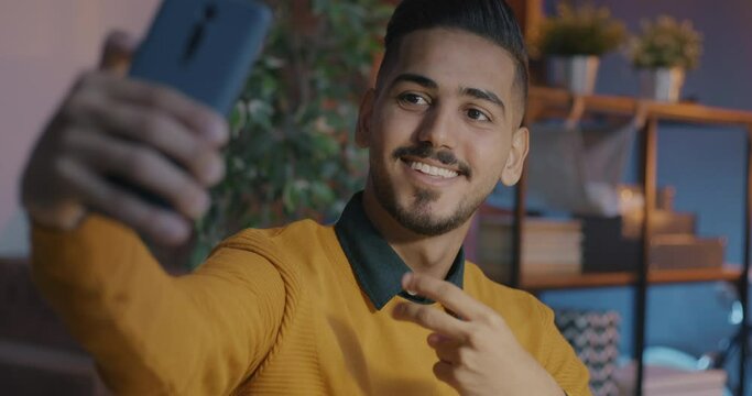 Slow motion of cheerful Middle Eastern man taking selfie posing for smartphone camera showing thumbs-up in office at night. Business and photo concept.