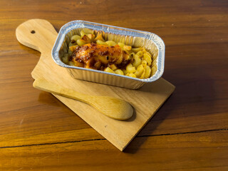 Selective focus of baked schotel macaroni in aluminum foil cup on wooden cutting board on wooden...