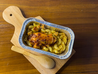 Selective focus of baked schotel macaroni in aluminum foil cup on wooden cutting board on wooden...
