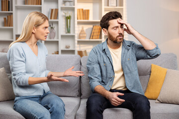 A woman, enraged, confronts her husband in their living space, he exhibits signs of a headache