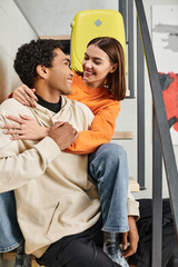 positive diverse couple sitting on stairs and smiling at each other during their trip, hostel room