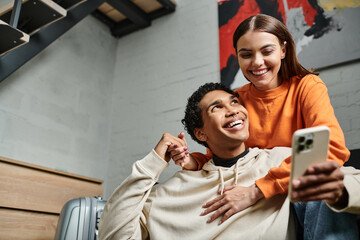 focus on happy diverse couple browsing internet from smartphone near luggage in hostel room