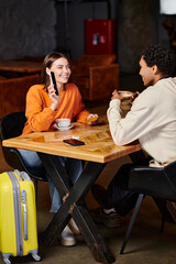 black man and woman using smartphone while sitting at table with coffee in cafe, luggage and travel