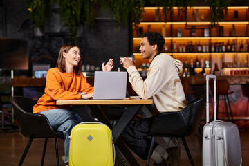 Fototapeta premium black man and woman laughing and talking at a cafe table, with their suitcases beside them, laptop