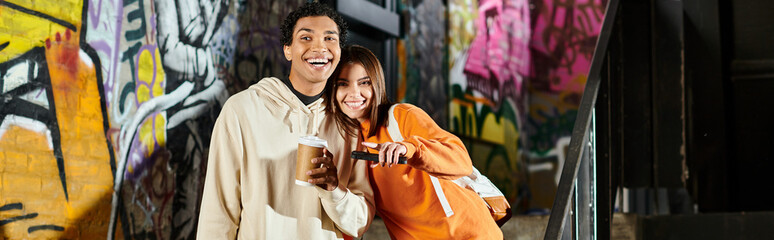Happy couple in casual wear with coffee cup and smartphone near graffiti painted wall, banner © LIGHTFIELD STUDIOS