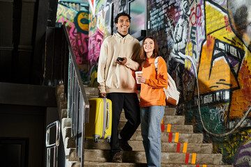 happy diverse couple standing on stairs with graffiti on wall, black man holding a yellow suitcase