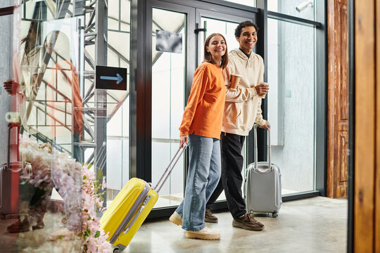happy diverse travel couple with suitcases chatting with coffee cups by glass doorway of hostel