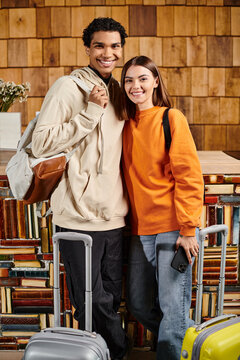 interracial happy couple standing with smartphone and luggage near book shelves in hostel, travel