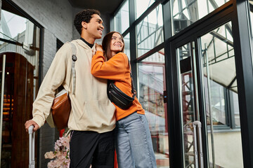 happy woman embracing black boyfriend near luggage in hostel, ready for journey as a couple together