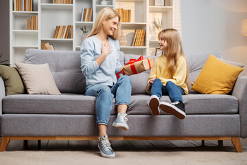 Celebrating Mother's Day, little girl surprises her mother with a gift box on their comfortable sofa