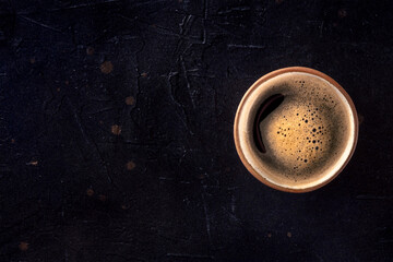 A cup of black coffee with froth, overhead flat lay shot on a black background, with copy space