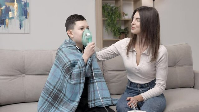 Young woman helping illness boy with nebulizer inhalation at home. A young mother helping her son to inhale with a nebulizer at home