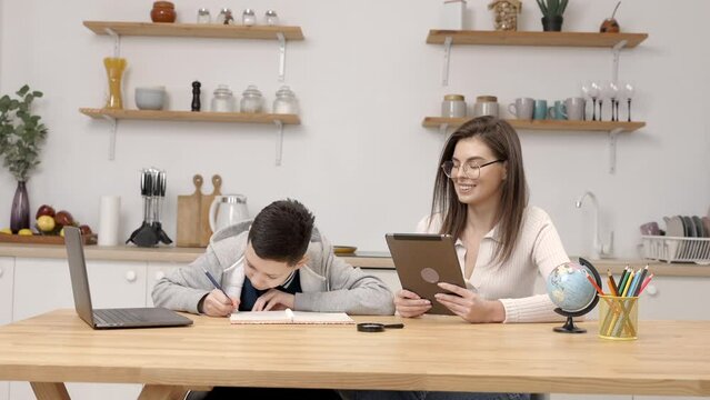 A young woman helps a boy with his lessons using a tablet and a globe. A homeschooling student does his homework with the help of a tutor