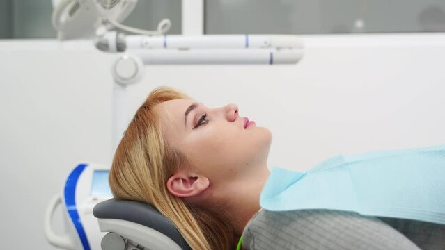 Female patient lying in dental chair in modern dental office. Preparing patient for UV teeth whitening. Blonde woman prepares for medical procedure in the private clinic. Dentist adjusts chair.