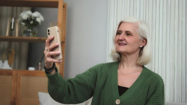 Happy smiling mature retired senior woman sitting on the sofa and making photo selfie, posing for photography, having video call conference. Joyful attractive Grandmother holding phone for call