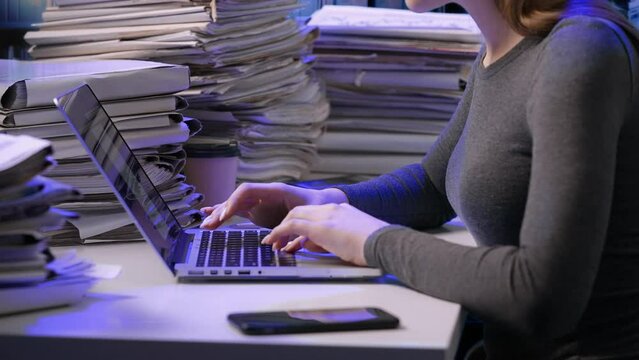 Exhausted young business woman office employee at the desk working late at night overloaded with paperwork. Female working on laptop, typing text inserting data.