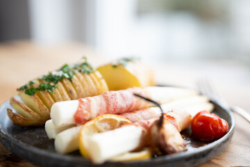Fried white asparagus wrapped in bacon with herbed potatoes and tomatoes on black ceramic plate and...