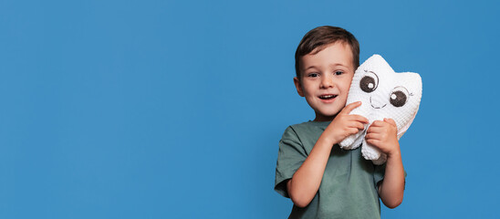 A smiling boy with healthy teeth holds a toothbrush and a plush tooth on a blue background. Pediatric dentistry. A place for your text. Panoramic photo.