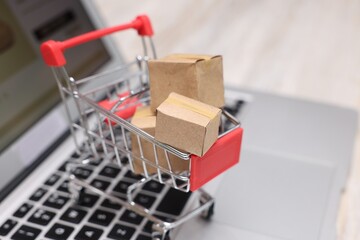 Internet store. Small cardboard boxes, shopping cart and laptop on light wooden table, closeup....