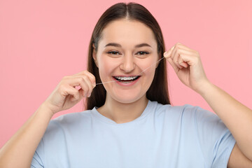 Smiling woman with braces cleaning teeth using dental floss on pink background