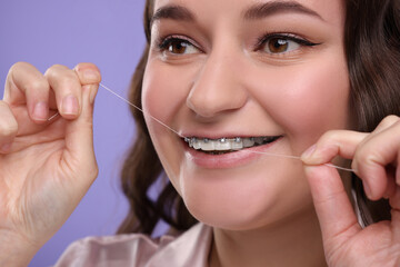 Woman with braces cleaning teeth using dental floss on violet background, closeup