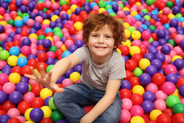 Happy little boy sitting on colorful balls in ball pit