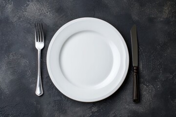 Top view of a white plate with a fork at the left side and a table knife at the right side on a dark grey backdrop.