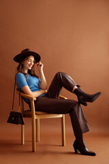 Fashionable happy smiling woman wearing hat, blue t-shirt, faux leather pants, pointed toe ankle cowboy boots, with trendy purse, posing on brown background. Full-length studio fashion portrait